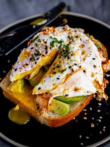 egg topped with chives and everything bagel seasoning on a piece of toast with avocado, the yolk is broken open so the inside is spilling out onto the plate.
