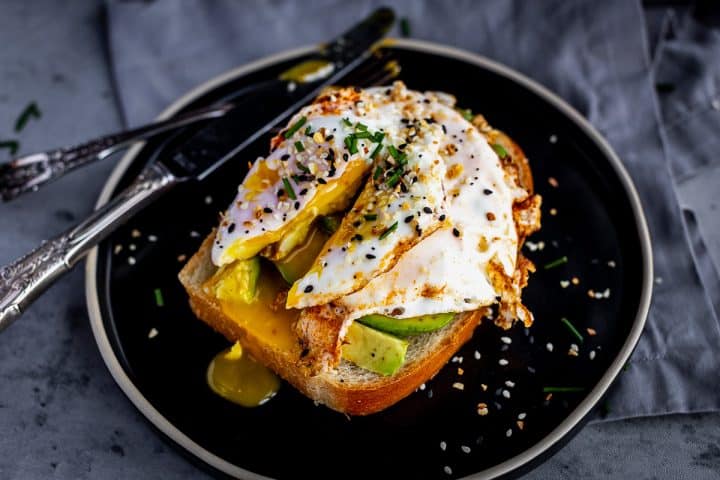 egg topped with chives and everything bagel seasoning on a piece of toast with avocado, the yolk is broken open so the inside is spilling out onto the plate.